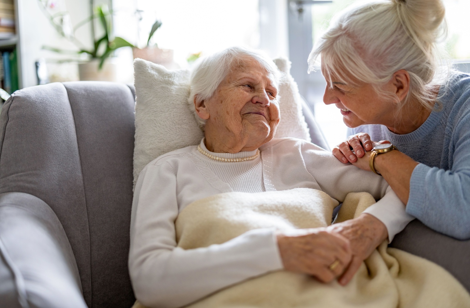 An adult child smiles and leans over their parent, who relaxes in an armchair in a memory care community.