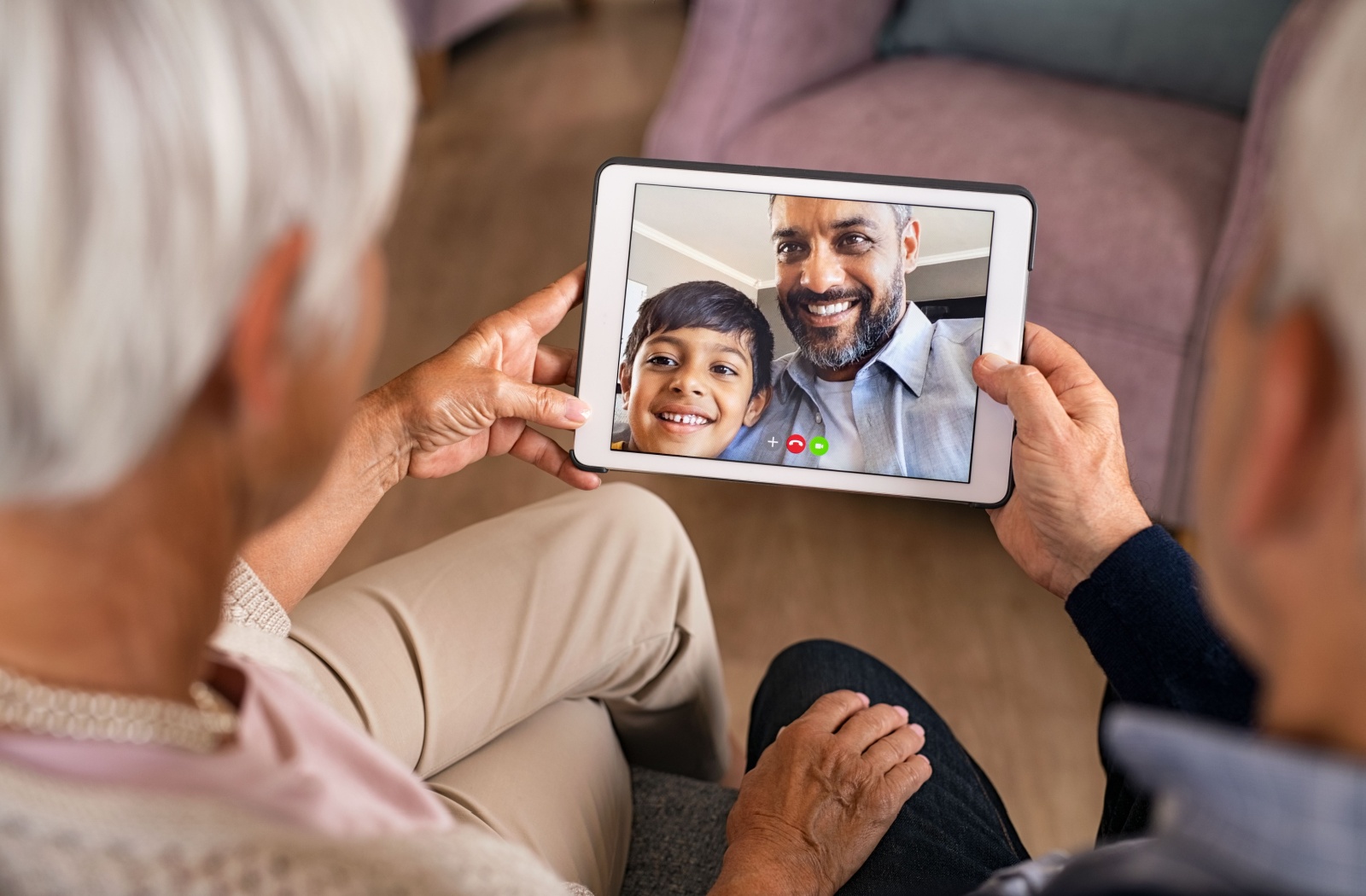 A senior couple each hold one end of a tablet to view a video call of their adult child and grandchild for a virtual visit