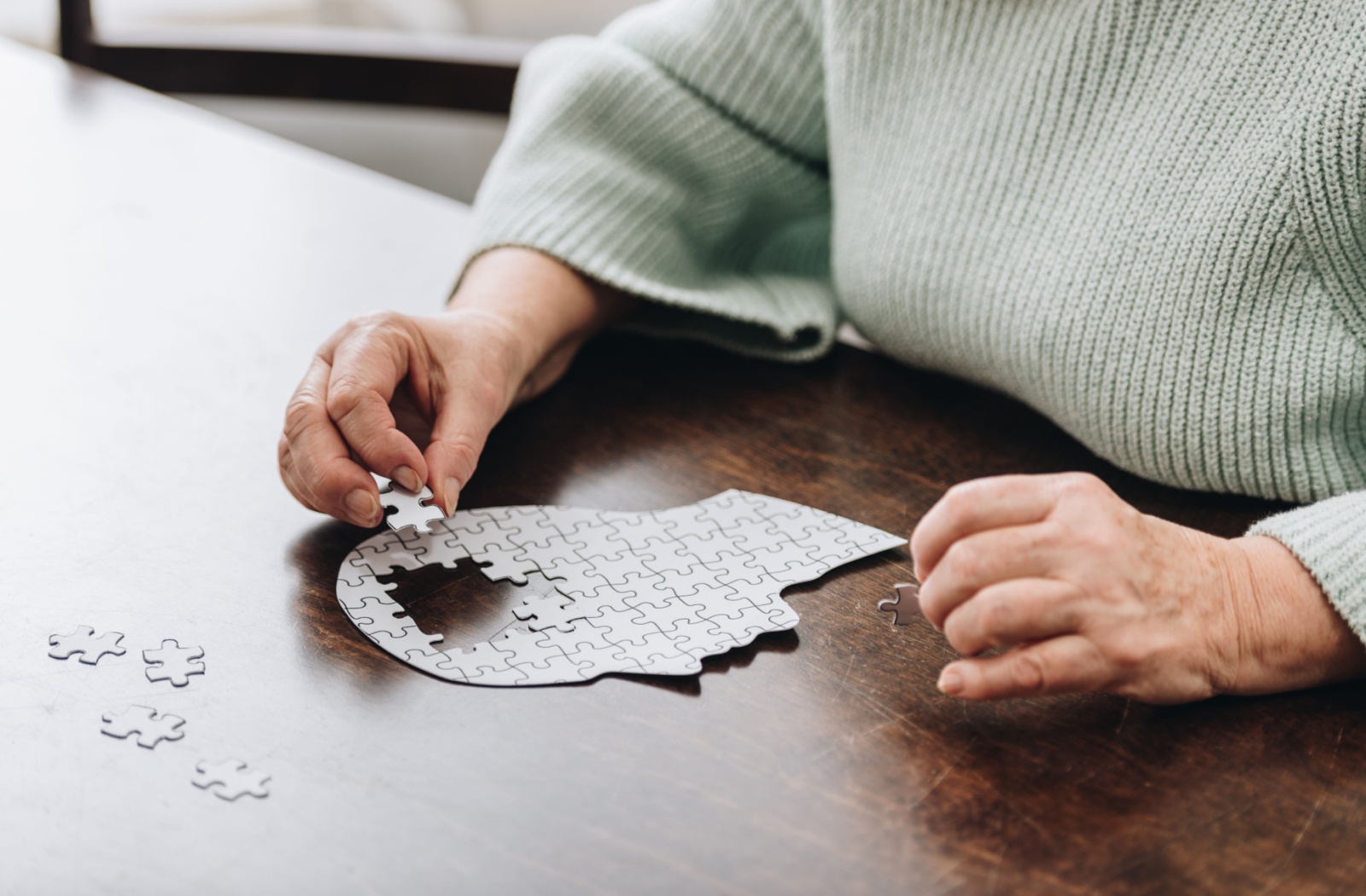 An older adult sits at a wooden table and assembles a white jigsaw puzzle of a head