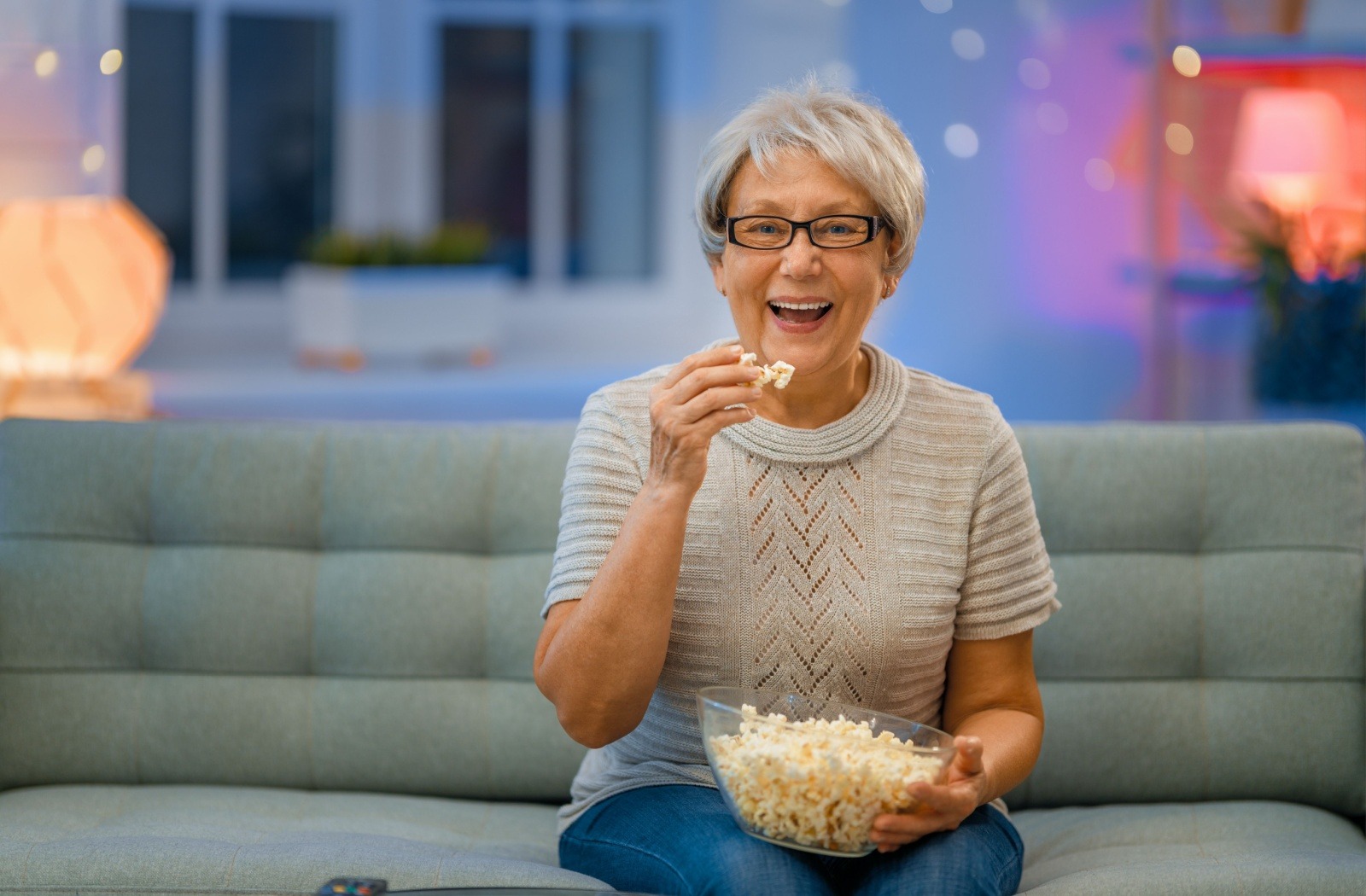 A smiling senior holds a bowl of popcorn while sitting on a gray couch.

