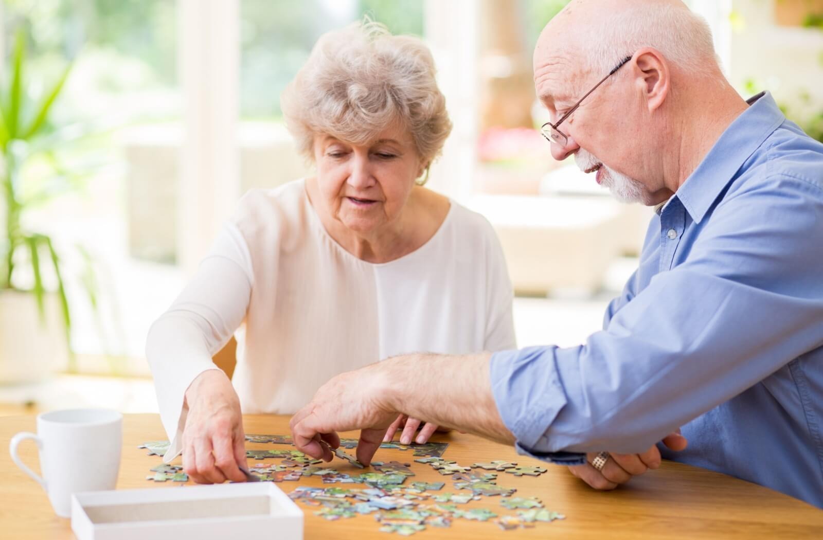 An older couple in a well-lit memory care common area work together to assemble a jigsaw puzzle
